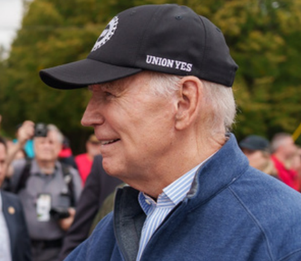 President Joe Biden is pictured outdoors, wearing a black baseball cap with the words "UNION YES" on the side. He is dressed in a blue jacket over a collared shirt. The background shows a blurred crowd of people and trees, indicating an event or gathering.